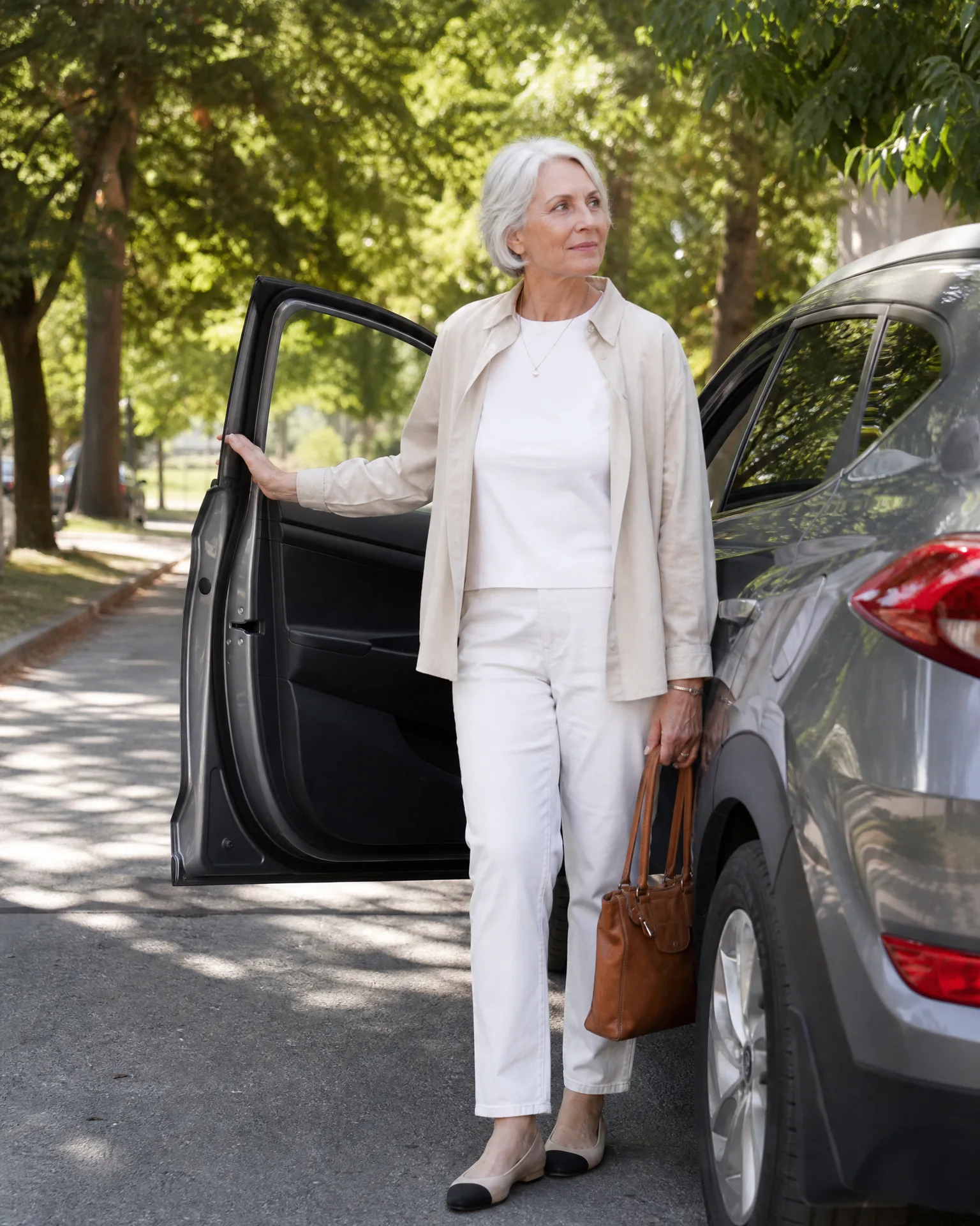 An older woman with short white hair, wearing a cream jacket, white top and trousers, stands holding a brown handbag beside an open car door of a grey SUV on a tree-lined street.