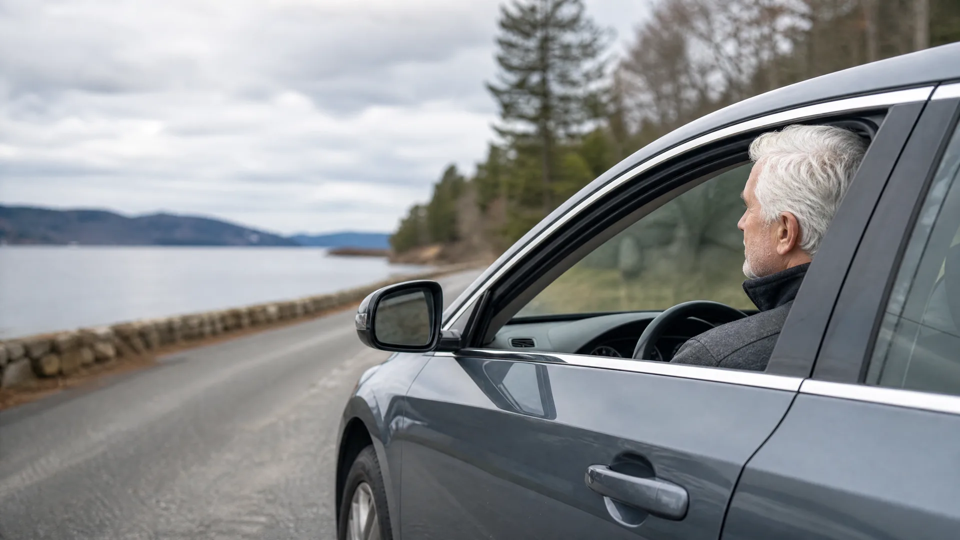 A gray-haired man sits in the driver's seat of a gray car with the window down, parked on a roadside beside a stone wall and a large body of water with low hills and conifers in the background under an overcast sky.