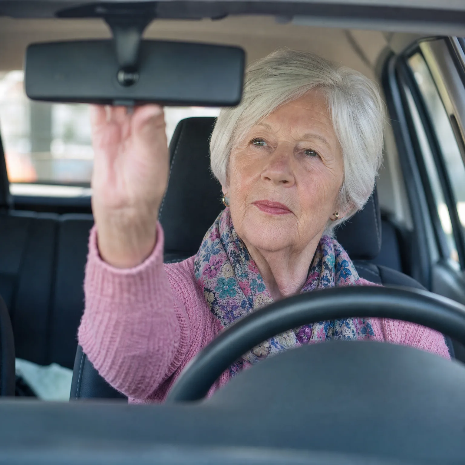 An older woman with short white hair, wearing a pink sweater and a floral scarf, sits in the driver's seat of a car and reaches up to adjust the rearview mirror.
