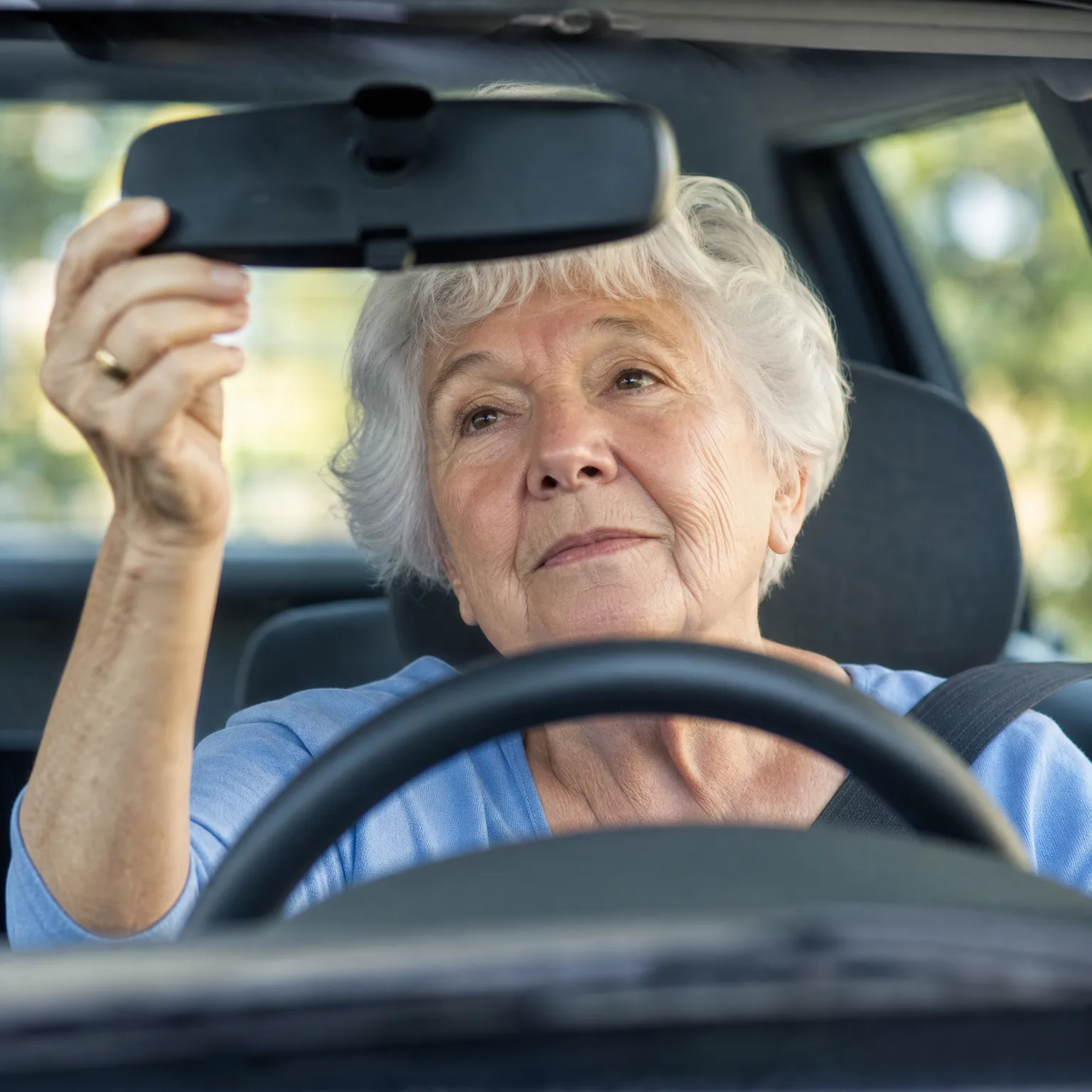 An older woman with short white hair, wearing a blue top and seat belt, sits in a car behind the steering wheel and reaches up with her right hand to adjust the rearview mirror, with green foliage blurred outside the windows.