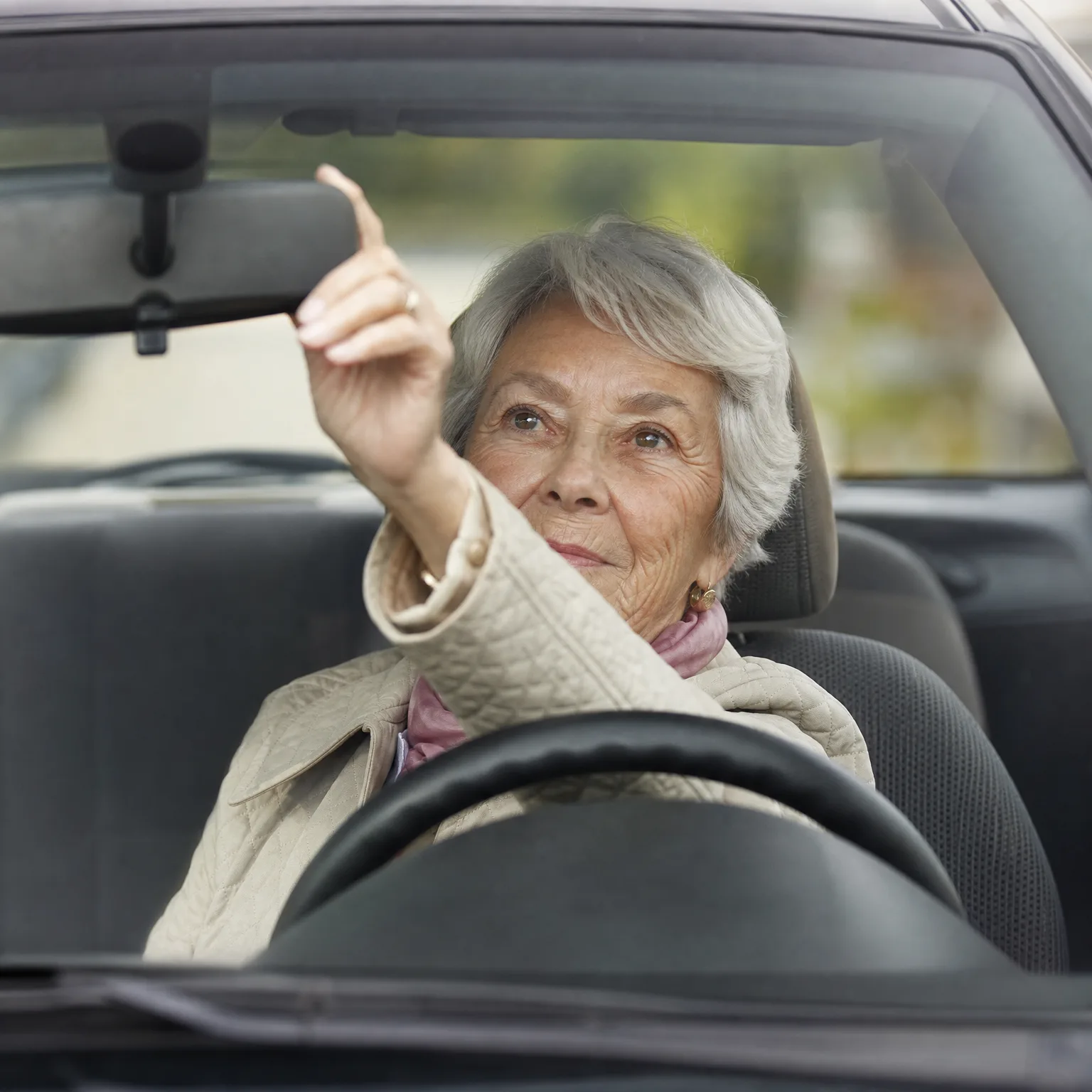 An older woman with short gray hair sits in the driver's seat of a car, seen through the windshield, raising one hand toward the rearview mirror area.