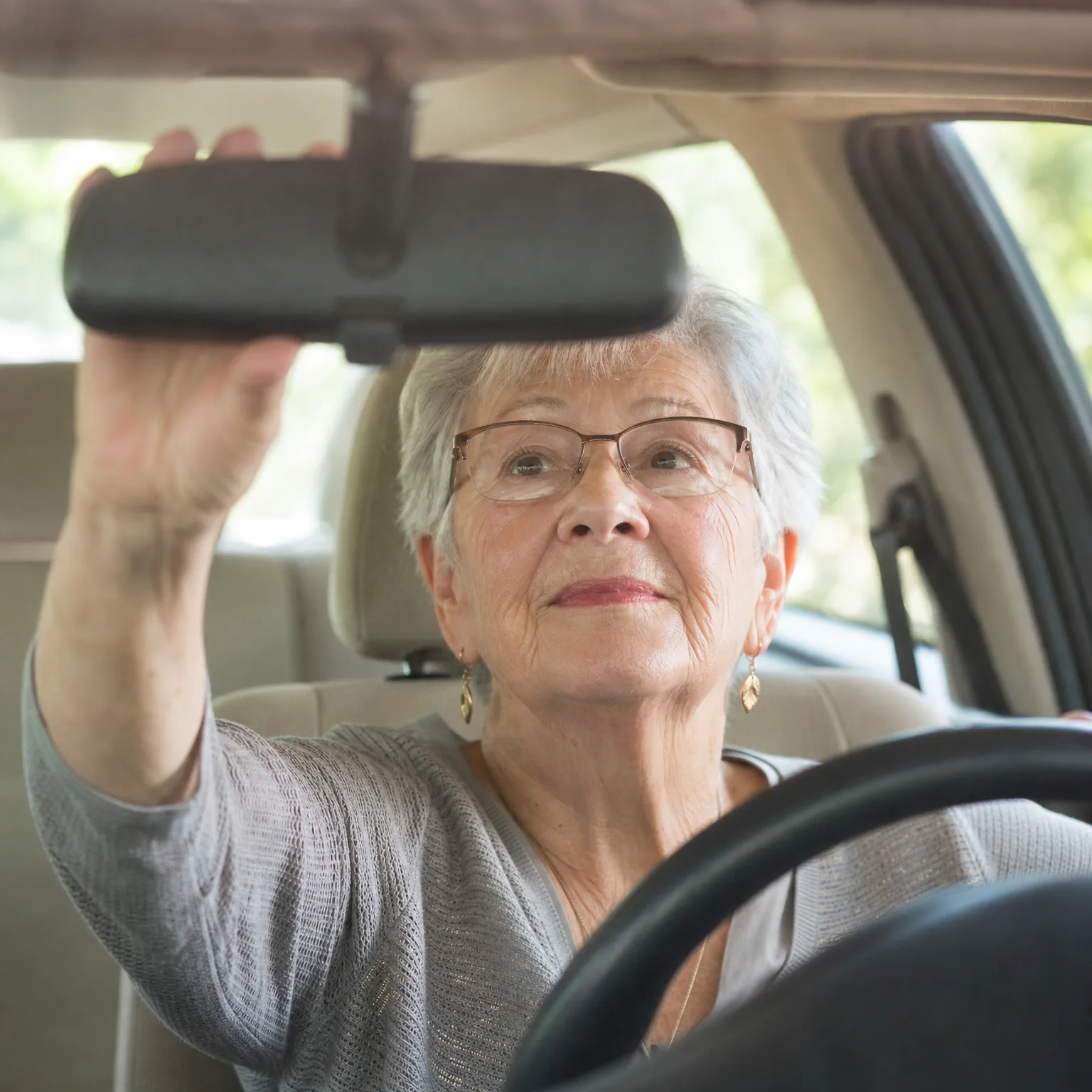 An older woman with short gray hair and glasses sits in a car's driver seat, reaching up with her right hand to adjust the rearview mirror, with the steering wheel in the foreground.