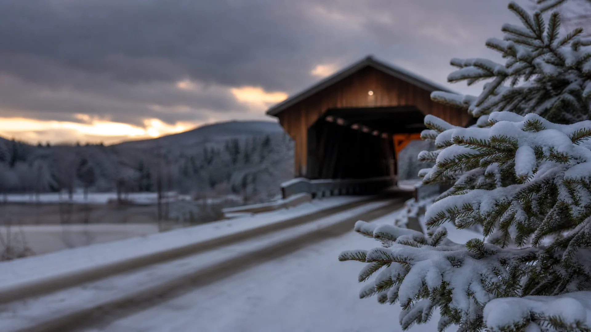A wooden covered bridge at the end of a snow-covered road at dusk, with a snow-laden spruce branch in the foreground and hills under a cloudy sky behind.